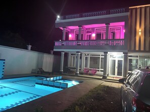 A two-story house with a swimming pool, a balcony with pink lighting, and a car parked nearby. - 5 BEDROOM MODERN  VILLA IN DIANI MOMBASA KENYA. 10 MINUTES WALK TO BEACH (Diani Beach)
