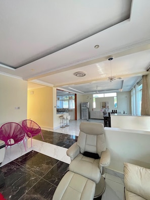 A spacious living area with a modern sofa, a dining table, and two vibrant chairs. A kitchen is visible in the background with a refrigerator and cabinets. - 5 BEDROOM MODERN  VILLA IN DIANI MOMBASA KENYA. 10 MINUTES WALK TO BEACH (Diani Beach)