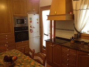 A kitchen with wooden cabinets, a microwave, a refrigerator, and a table with fruit. - Holiday Home 'Els Pins' with Private Terrace, Wi-Fi and Air Conditioning (Port d'Alcúdia)