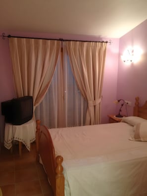 A bedroom with a bed, curtains, a television on a table, a bedside lamp, and a wall-mounted light fixture. - Holiday Home 'Els Pins' with Private Terrace, Wi-Fi and Air Conditioning (Port d'Alcúdia)