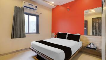 A hotel room with a bed, a window with curtains, an air conditioning unit, a ceiling fan, and a nightstand with a kettle.