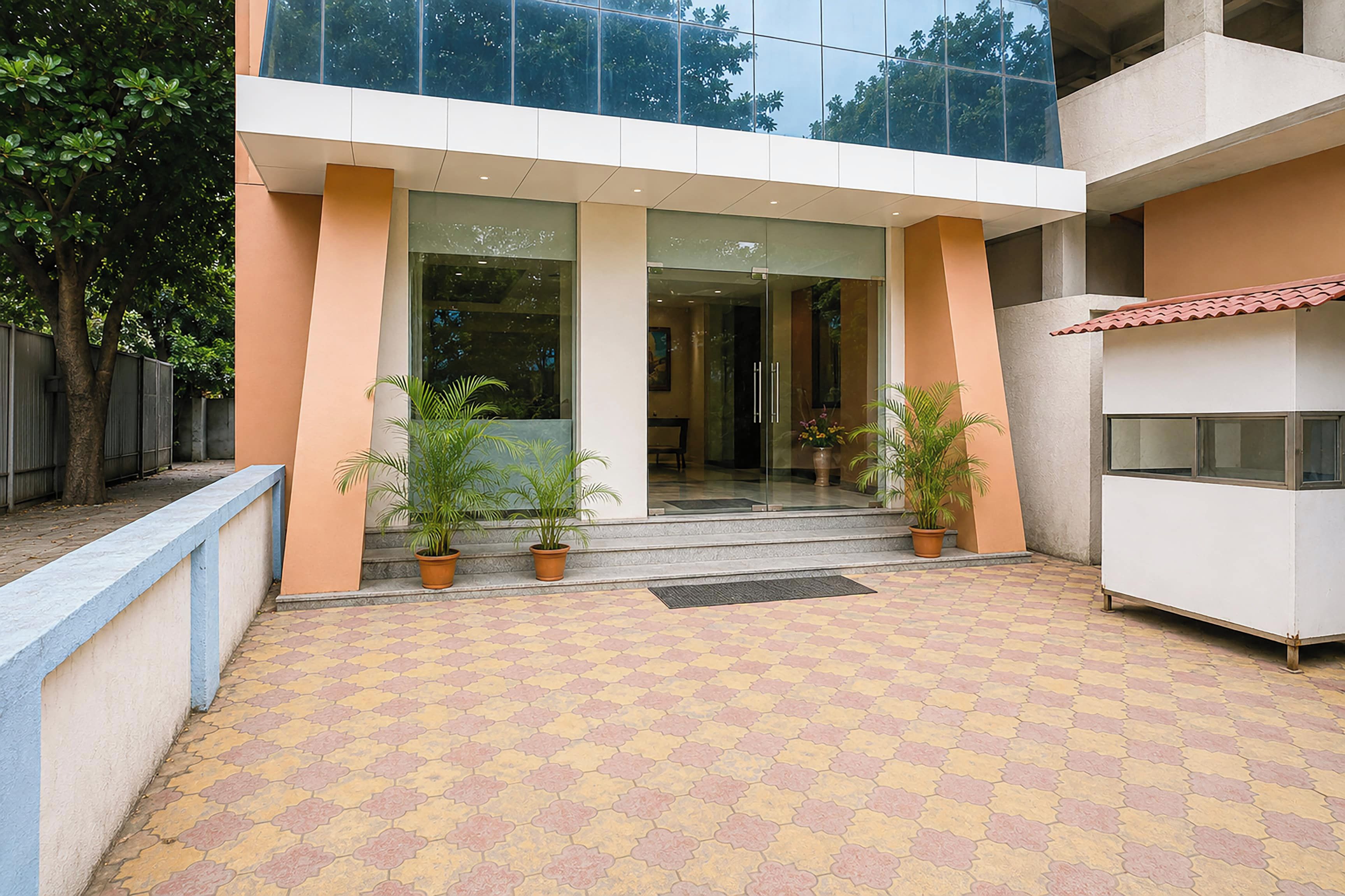 A building entrance with a tiled patio, potted plants, and a glass door leading inside.