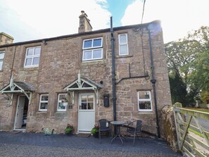 A stone building with a white door, two windows, and a small outdoor seating area with a table and chairs. - The Gables (Alnwick)