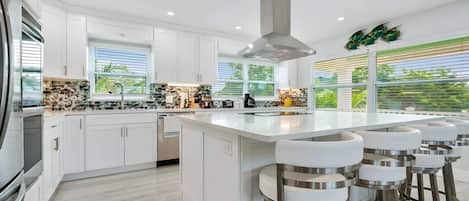 A modern kitchen with white cabinetry, a central island with bar stools, and a large window with blinds.