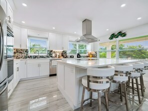 A modern kitchen with white cabinetry, a central island with bar stools, and a large window with blinds. - Two-story 3BR With Pool Table and Private Yard (St. Petersburg)