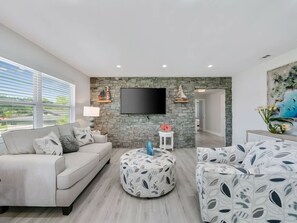 A modern living room with a stone wall, a flat-screen TV, a sofa, an ottoman, and a painting. - Two-story 3BR With Pool Table and Private Yard (St. Petersburg)
