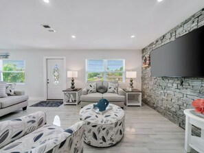A modern living room with a sofa, ottoman, and side tables, a large TV, and a stone accent wall. - Two-story 3BR With Pool Table and Private Yard (St. Petersburg)