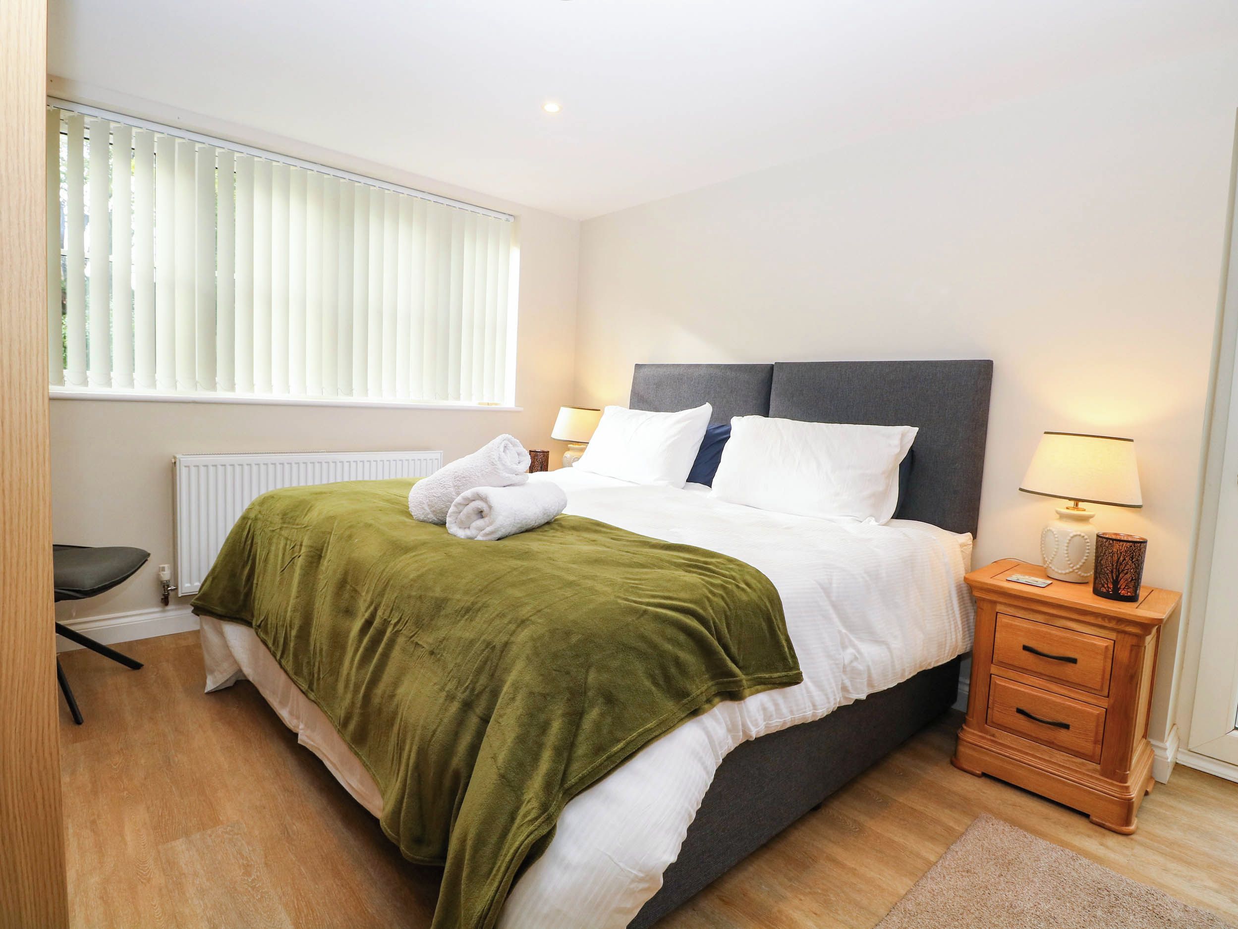 A neatly made bed with a green blanket, a bedside table with a lamp, and a chair.