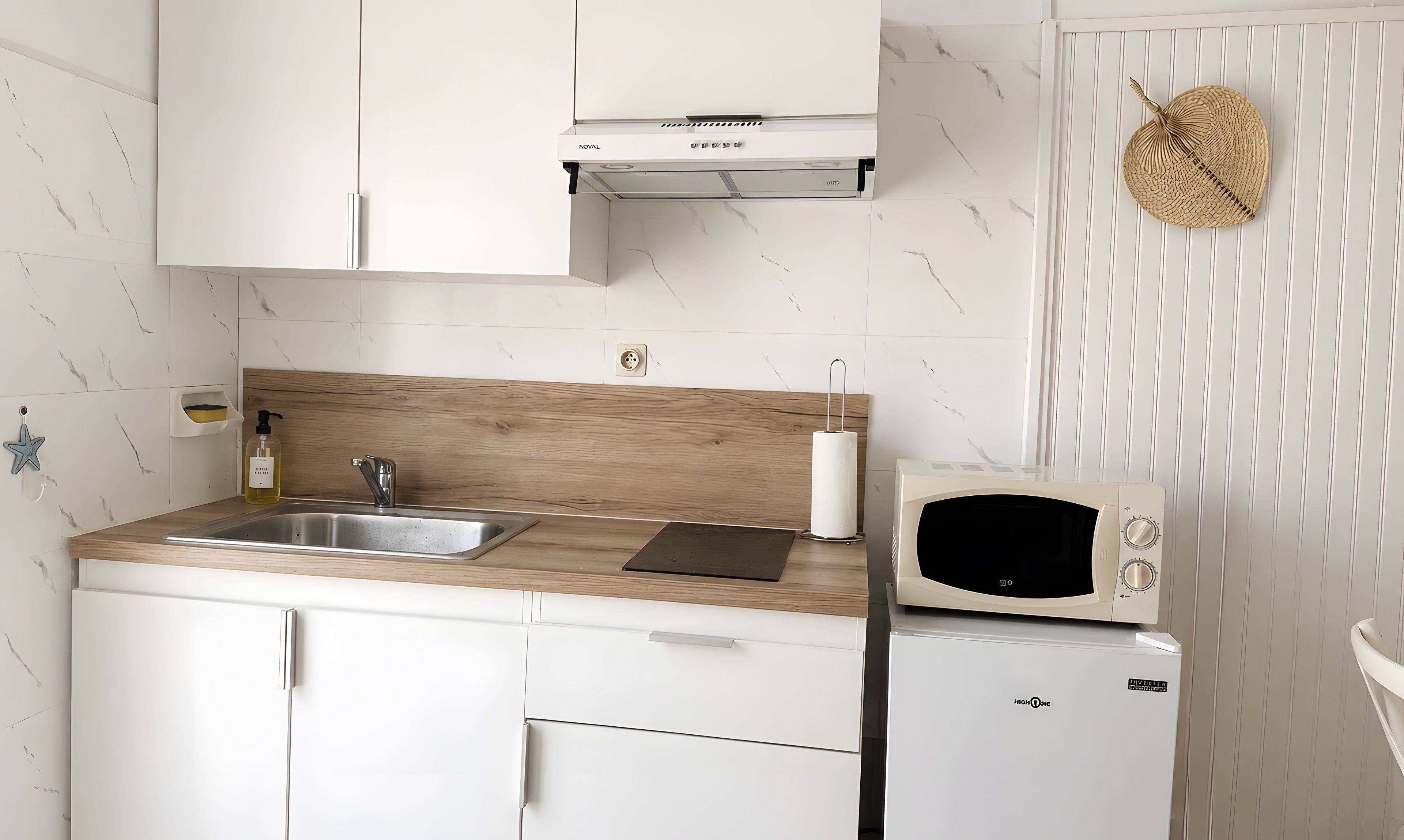 Une cuisine avec des meubles blancs, un plan de travail en bois, un évier, un four à micro-ondes et un petit réfrigérateur.