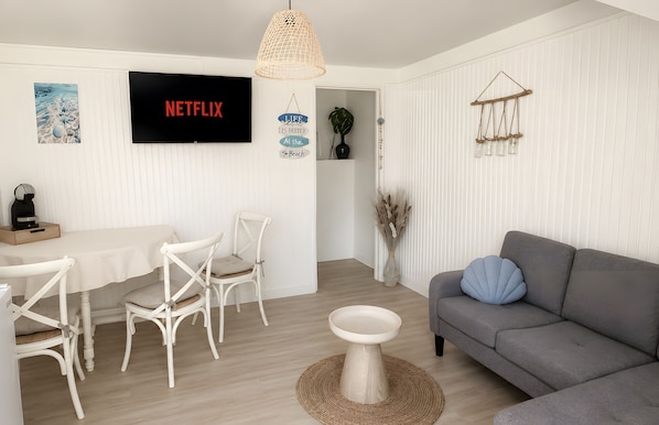A modern living room with a grey sofa, white chairs, a dining table, a TV mounted on the wall, and a hanging light fixture.