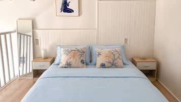 Une chambre à coucher comprenant un lit, deux tables de chevet, une commode, une lampe fixée au mur, un tableau encadré et une fenêtre donnant sur un paysage.