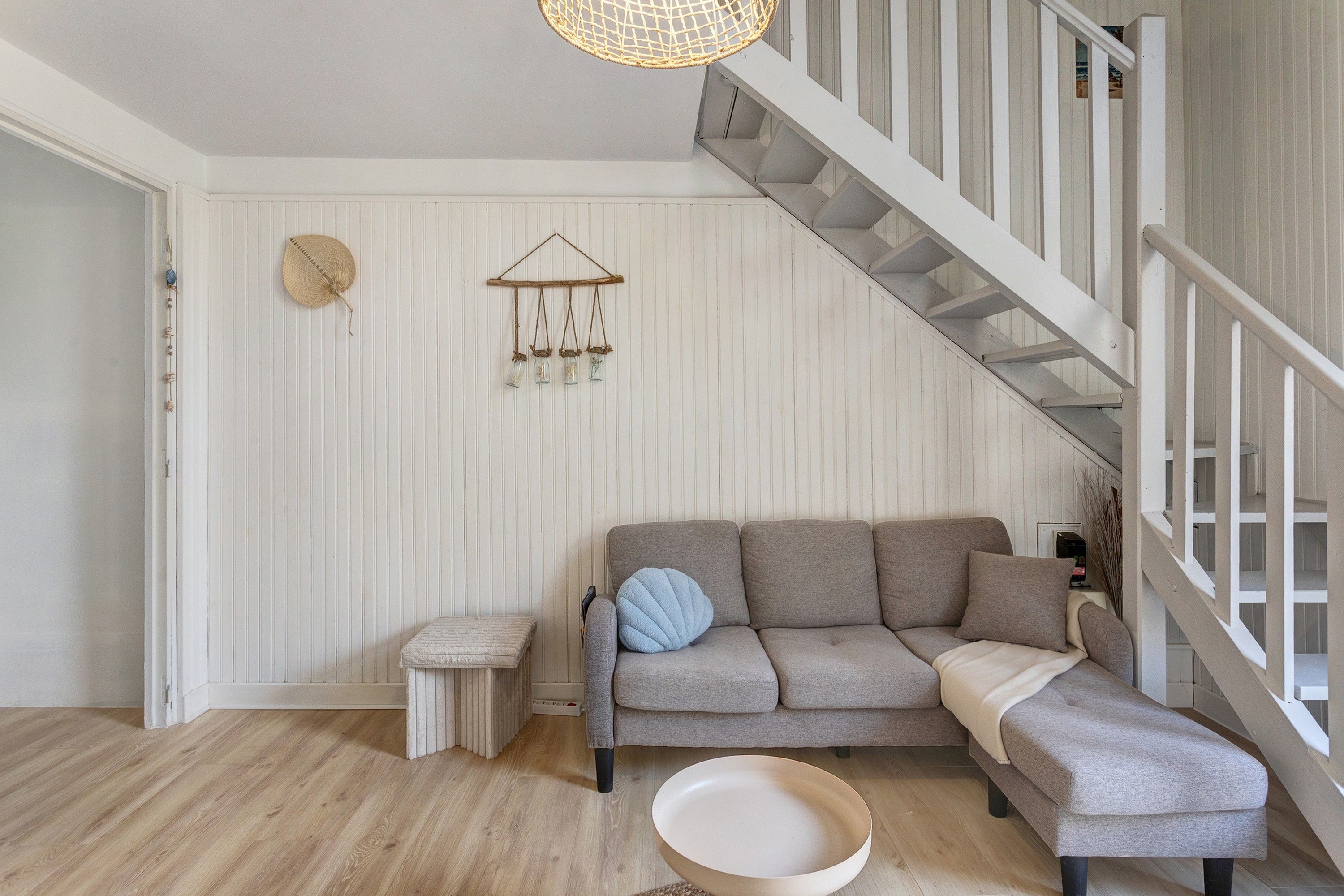 Ein Wohnzimmer mit grauem Ecksofa, einem kleinen runden Tisch, einer Pendelleuchte und einer Treppe.