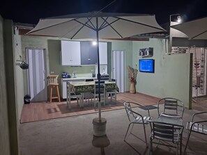A dining area with a table and chairs, a large umbrella, a kitchen area with cabinets, and a television on the wall.
