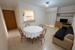 A living room with a round table and four chairs, a sofa, a television, and a kitchen area with cabinets and a window. - Residencial Ibis (Bauru)
