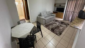 Una sala de estar con un sofá gris, una mesa de comedor con sillas, un ventilador de techo y una zona de cocina con un televisor.