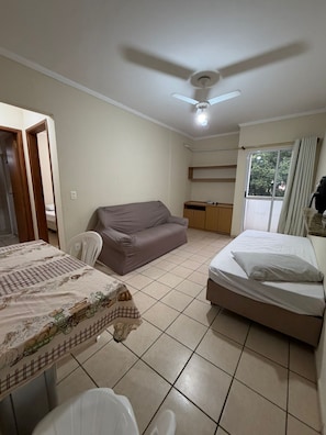 Ein Zimmer mit einem Bett, einem Schlafsofa, einem Esstisch mit Stühlen, einem Deckenventilator und einem Fenster mit Vorhängen.