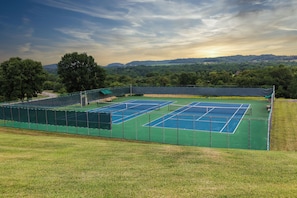Ein Tennisplatz mit mehreren Spielfeldern, umgeben von einem grünen Zaun, und eine Aussicht auf eine hügelige Landschaft im Hintergrund.