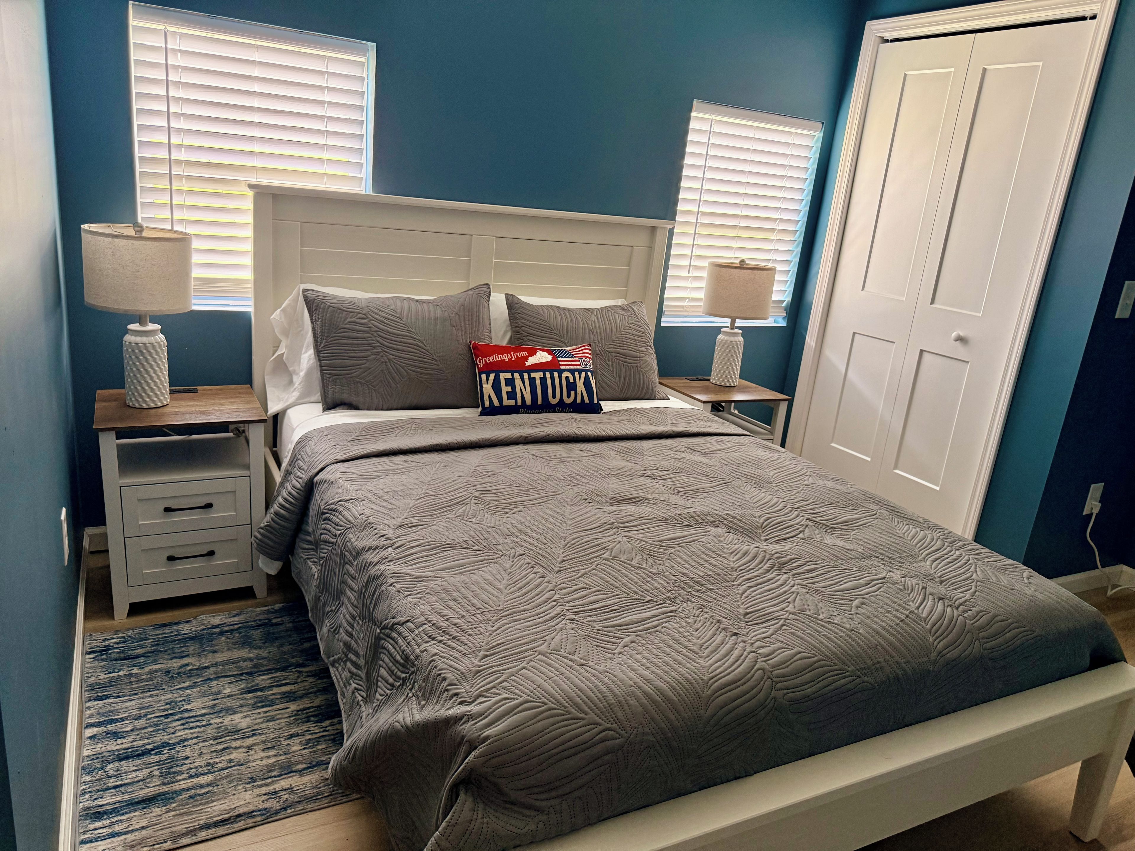 Een slaapkamer met een bed, twee nachtkastjes, een lamp op elk en een blauwe wand, plus een deur.