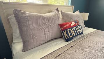 A bed with a quilted headboard, a pillow with 'Greetings from Kentucky' text, and a window with blinds.