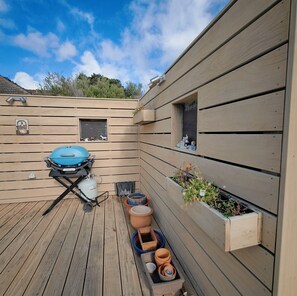Eine Holzterrasse mit einem Grill, einem Fenster mit Blick auf Bäume und einem Blumenkasten mit Pflanzen.