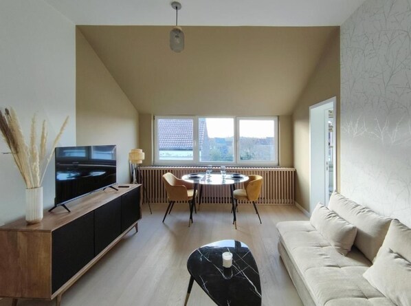 A modern living room with a sofa, a coffee table, a TV on a cabinet, a dining table with chairs, and a window with a view.