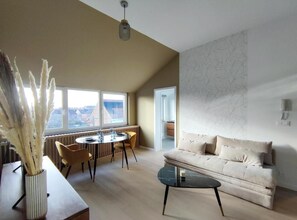 A modern living room with a sofa, a coffee table, a dining table with chairs, and a large window offering a view.