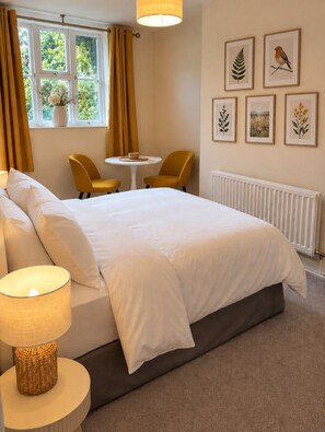 Hotelkamer met een bed, twee stoelen, een klein tafeltje en een raam met gordijnen. Aan de muur hangen ingelijste botanische prints.