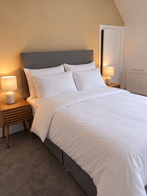 Een net opgemaakt bed met witte lakens, een grijs hoofdbord en twee bedlampjes op houten nachtkastjes.