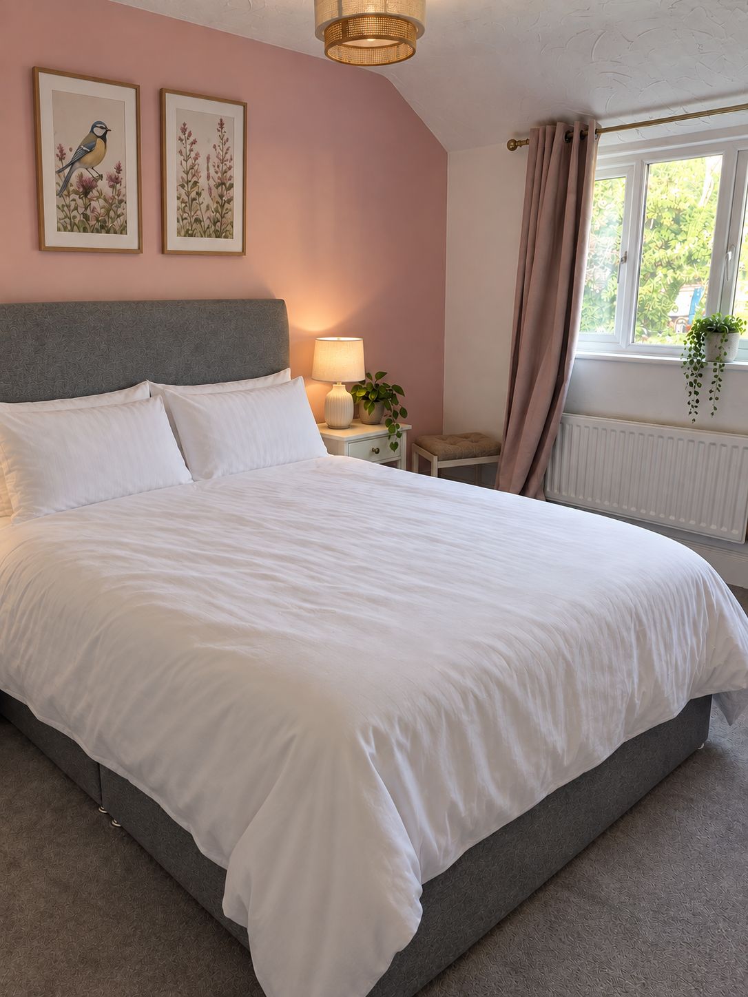 A bedroom with a large bed, a bedside table with a lamp, a window with curtains, and two wall-mounted pictures.