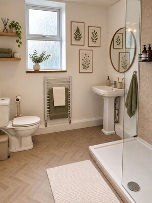 Een badkamer met toilet, wastafel en douche. Op de muur hangen botanische prints in lijsten, er is een radiator en een raam met een plant.