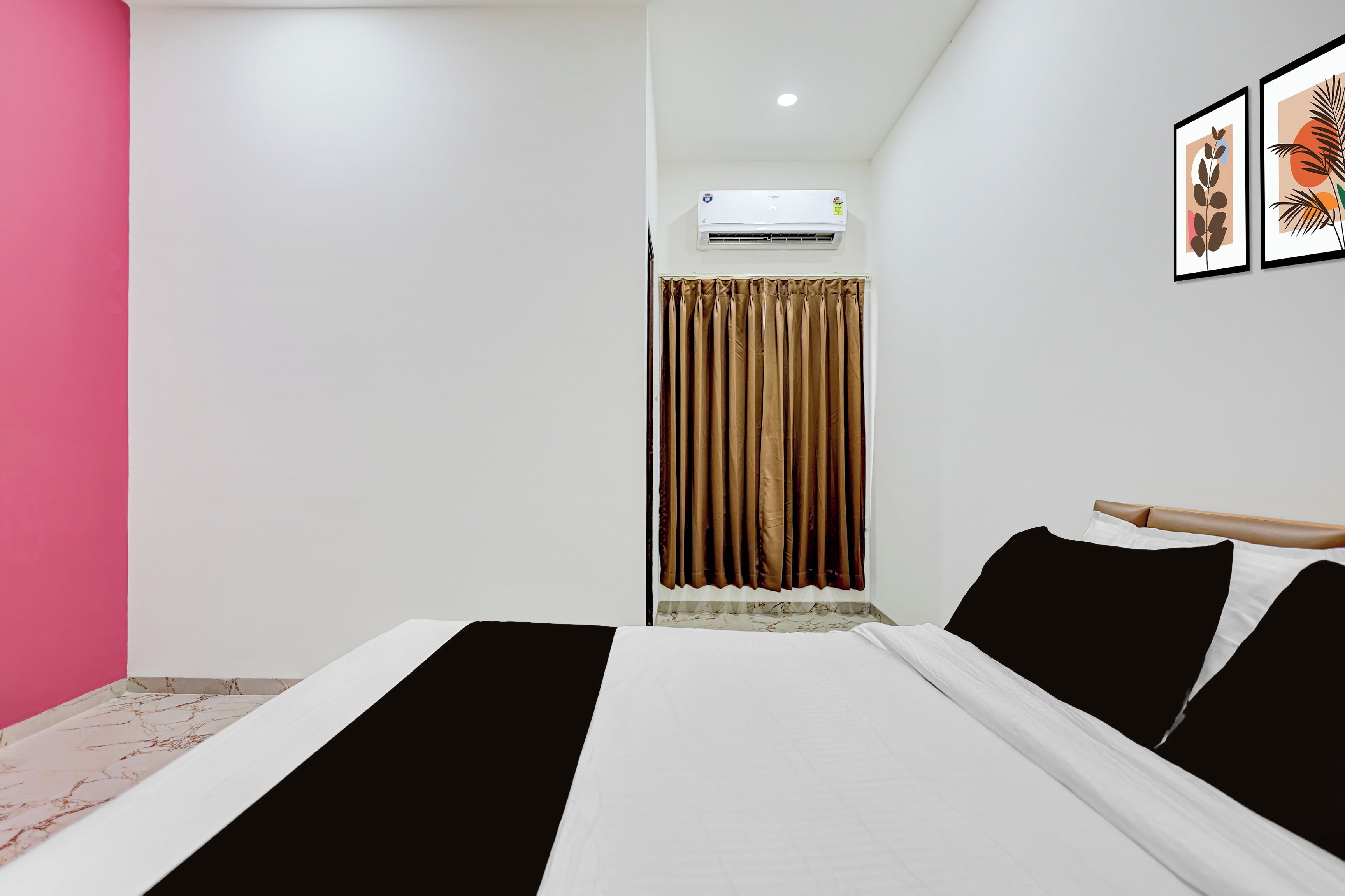 Una habitación de hotel con cama, ventana con cortinas, equipo de aire acondicionado y una pared decorada con obras enmarcadas.