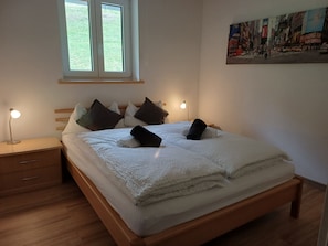 A neatly made bed with pillows, a bedside lamp, a wooden nightstand with a drawer, and a window with a view of greenery.