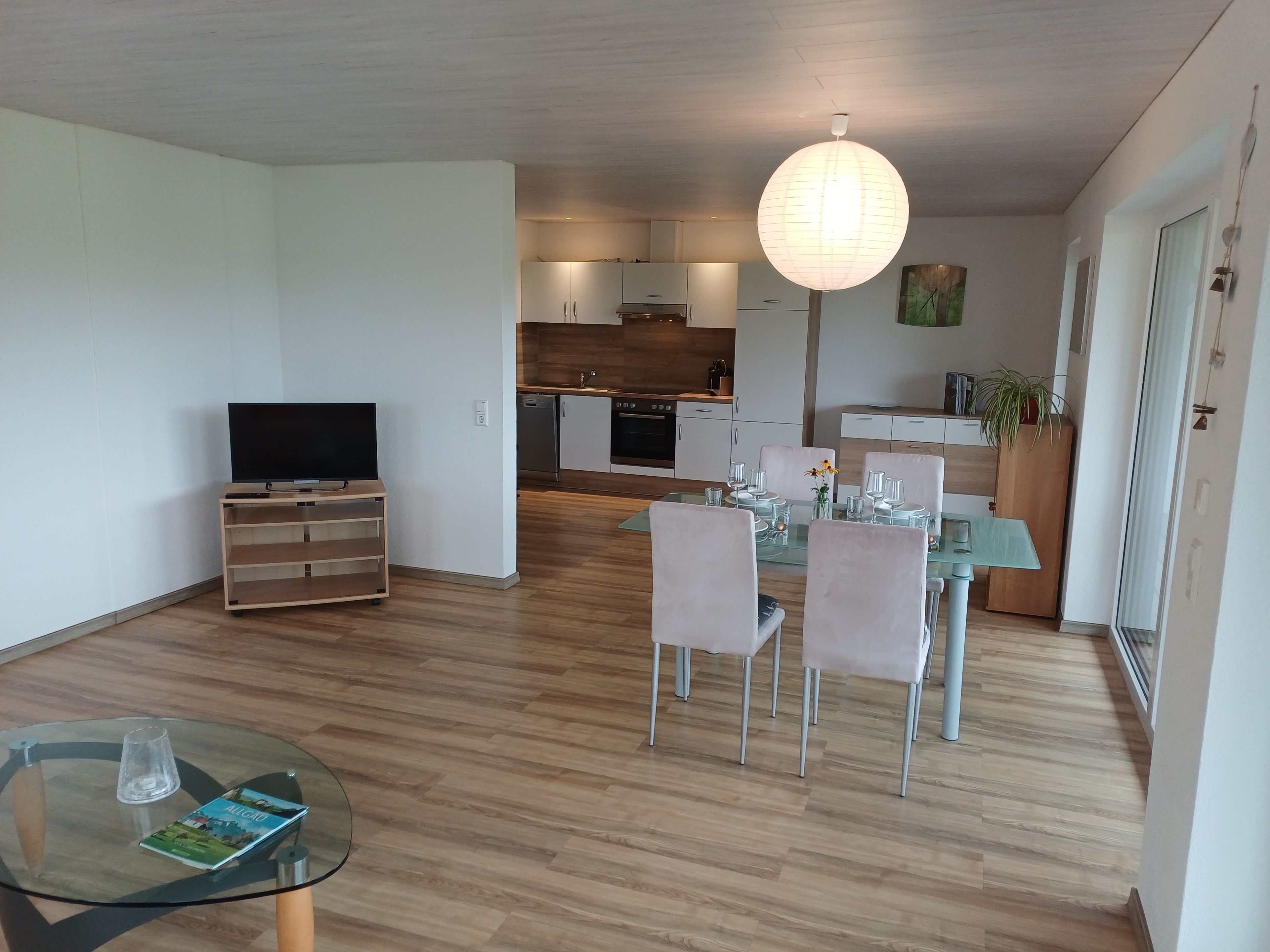 A modern living room with a glass dining table, a television on a wooden stand, and a kitchen in the background.