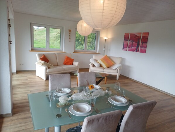 A dining area with a glass table set for six, surrounded by two sofas with cushions, a floor lamp, and a view of a green landscape through the windows.