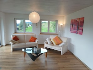 Un salón con dos sofás blancos, una mesa de centro de cristal y un ventanal con vistas a zonas verdes.