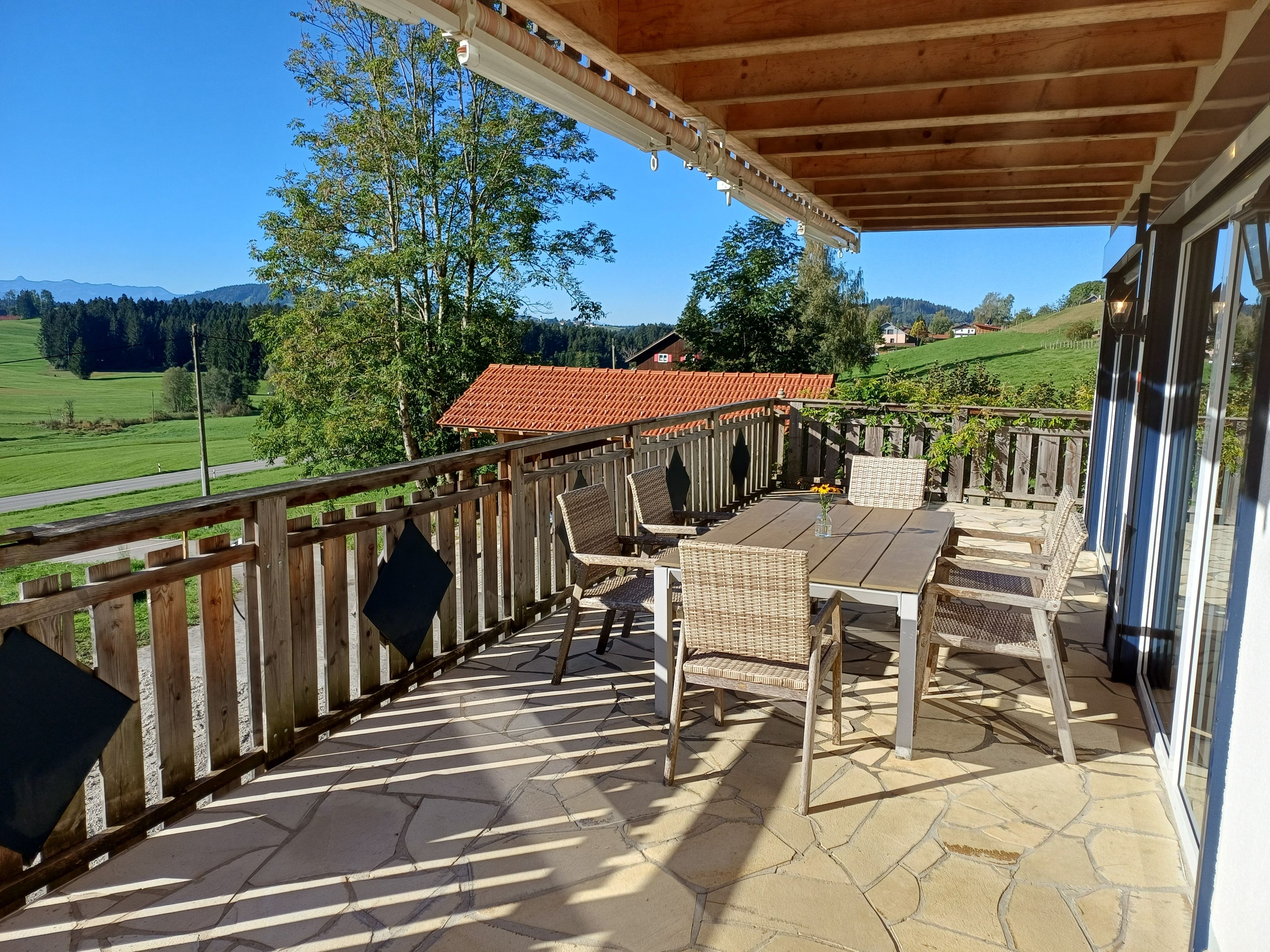 A covered patio with a table and chairs, a wooden railing, and a view of a red-roofed building and green landscape.