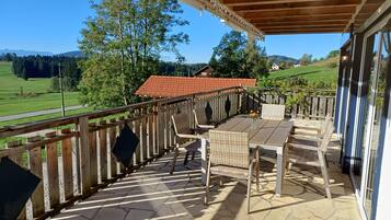 A covered patio with a table and chairs, a wooden railing, and a view of a red-roofed building and green landscape.