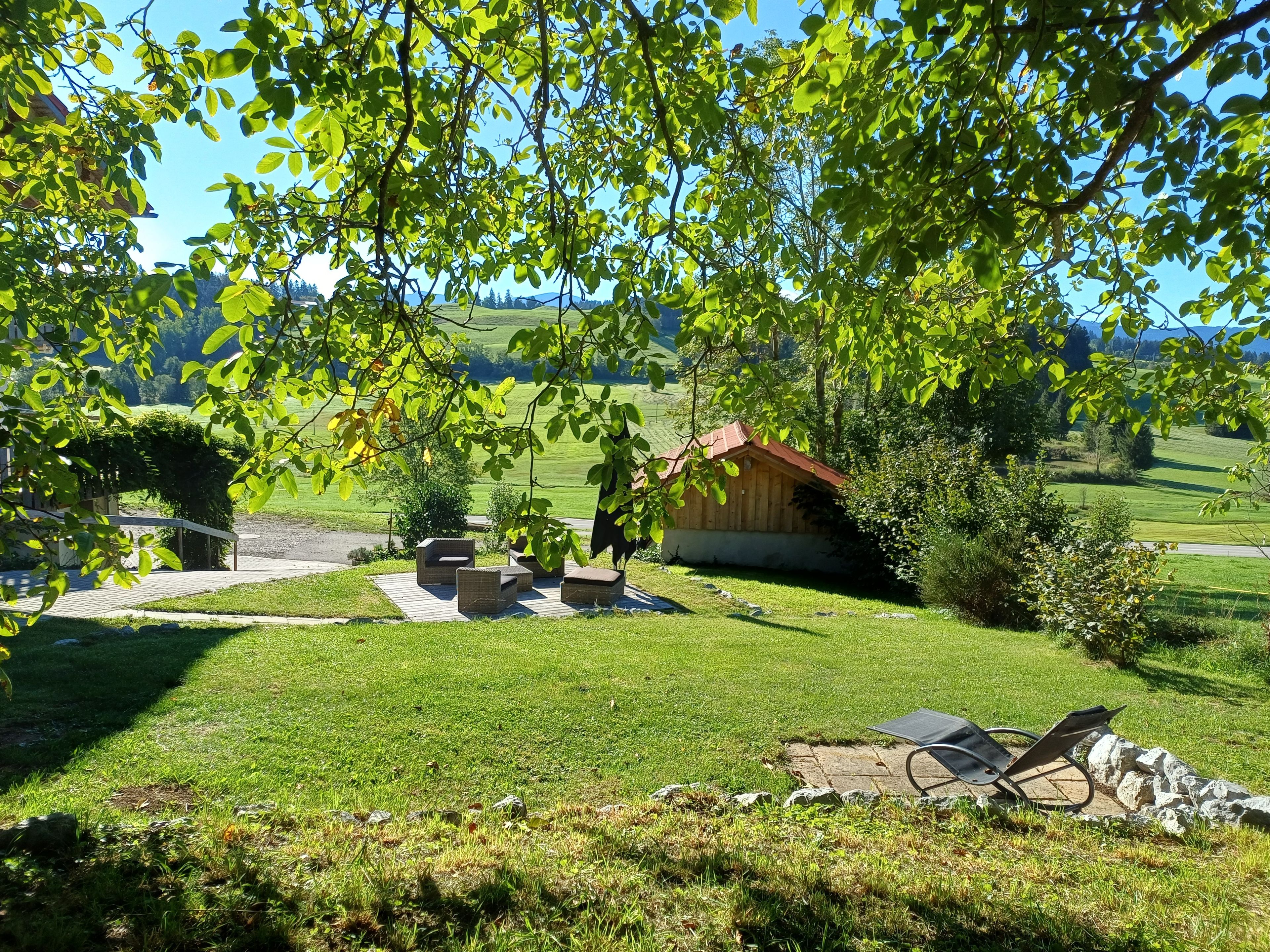 Eine grüne Außengelände mit einer Holzkonstruktion, Sitzgelegenheiten im Freien und Blick auf eine hügelige Landschaft.