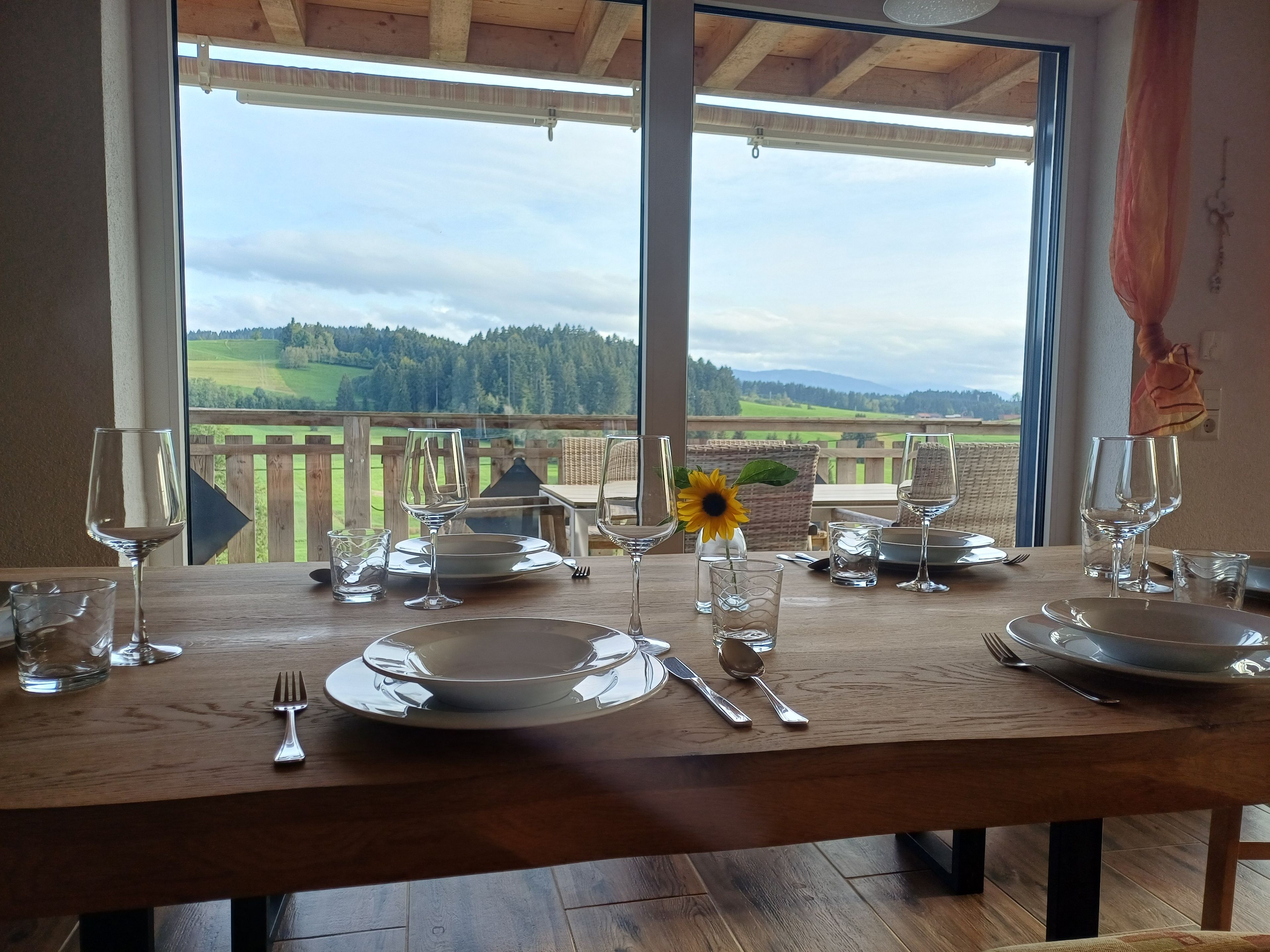 A dining table set for four with a view of a wooden deck and a forested landscape.