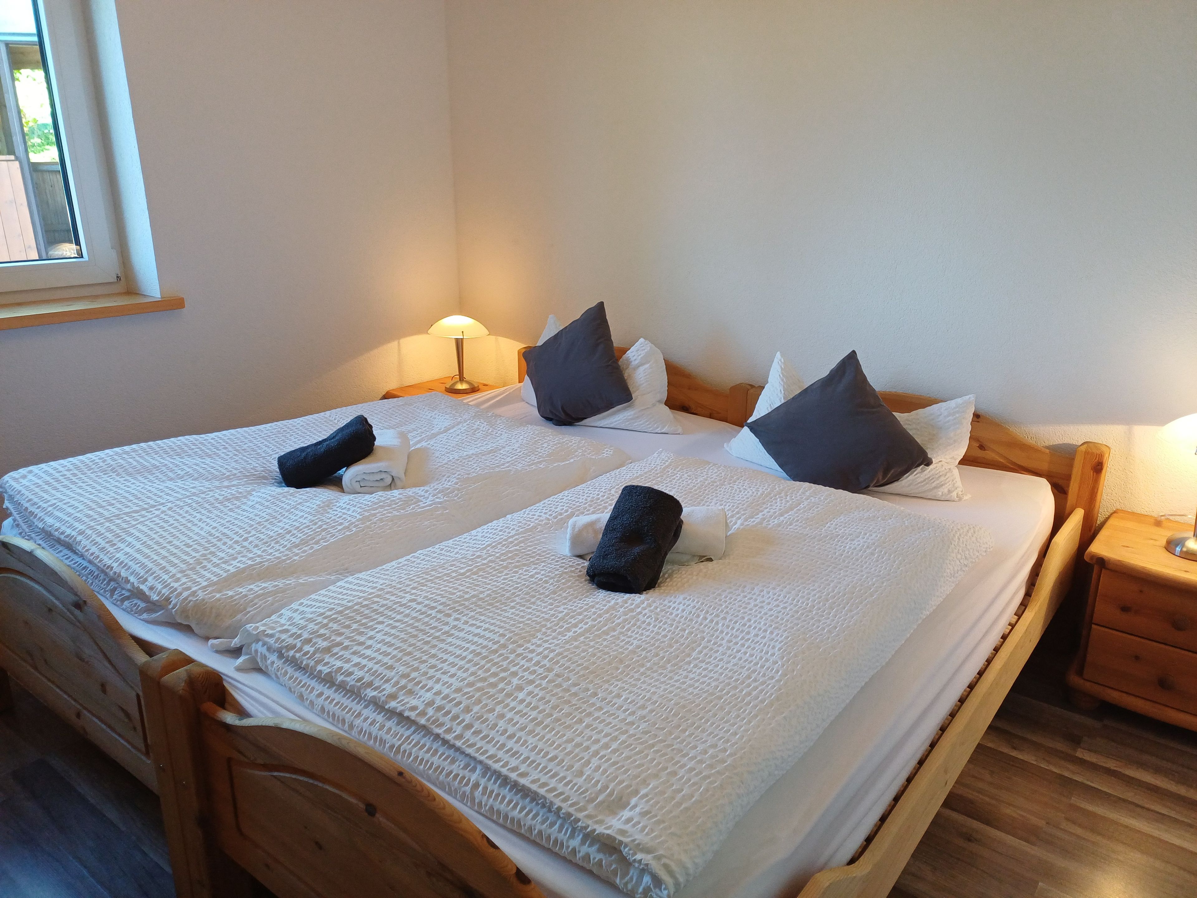 A double bed with a white textured bedspread, two pillows, a folded towel, and a bedside table with a lamp.