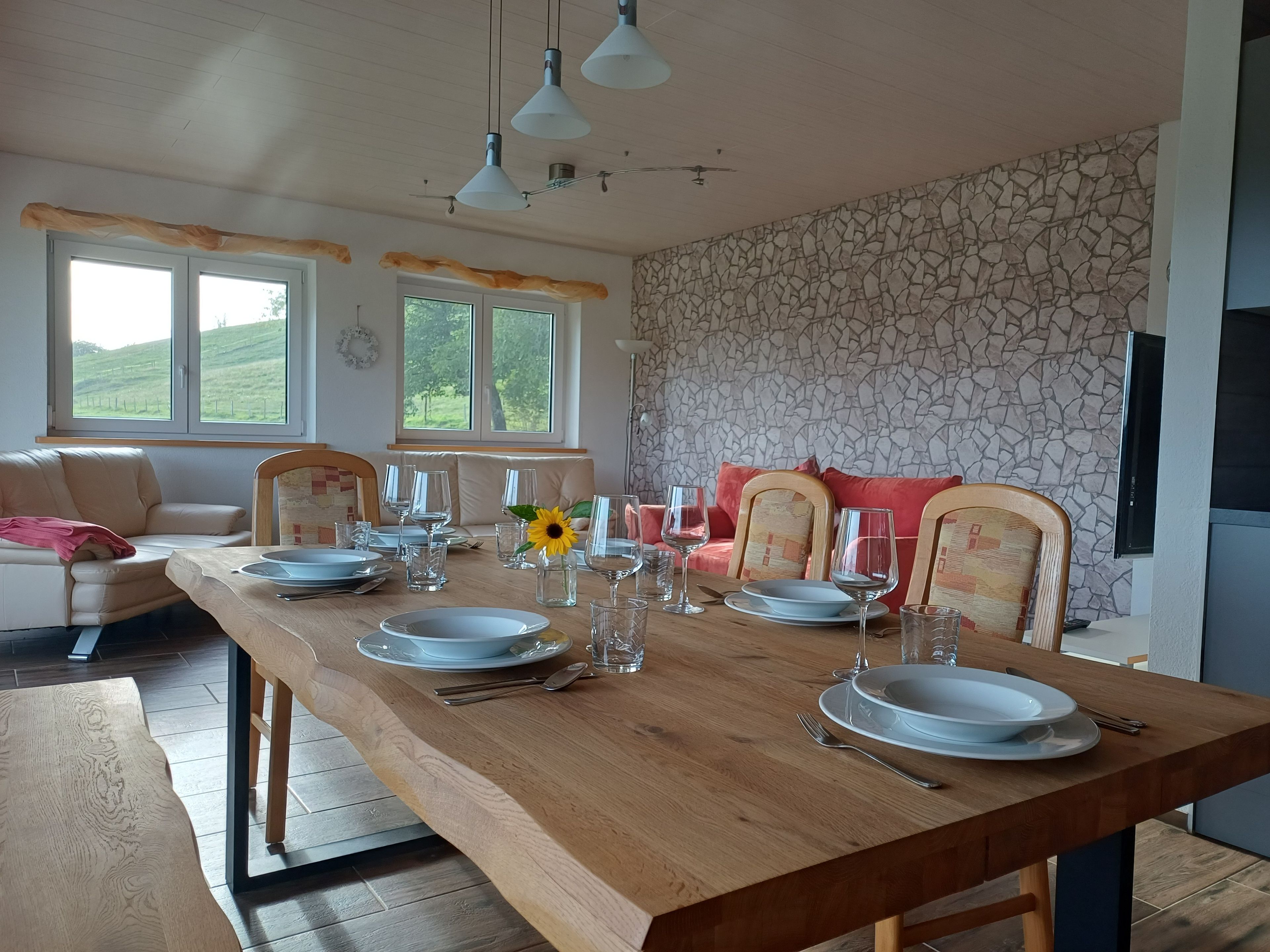 A dining area with a wooden table set for four, surrounded by cushioned chairs, a stone wall, and a view of the outdoors.