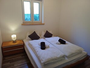 A neatly made bed with white linens and two pillows, a bedside lamp on a wooden nightstand, and a window with a view of greenery.