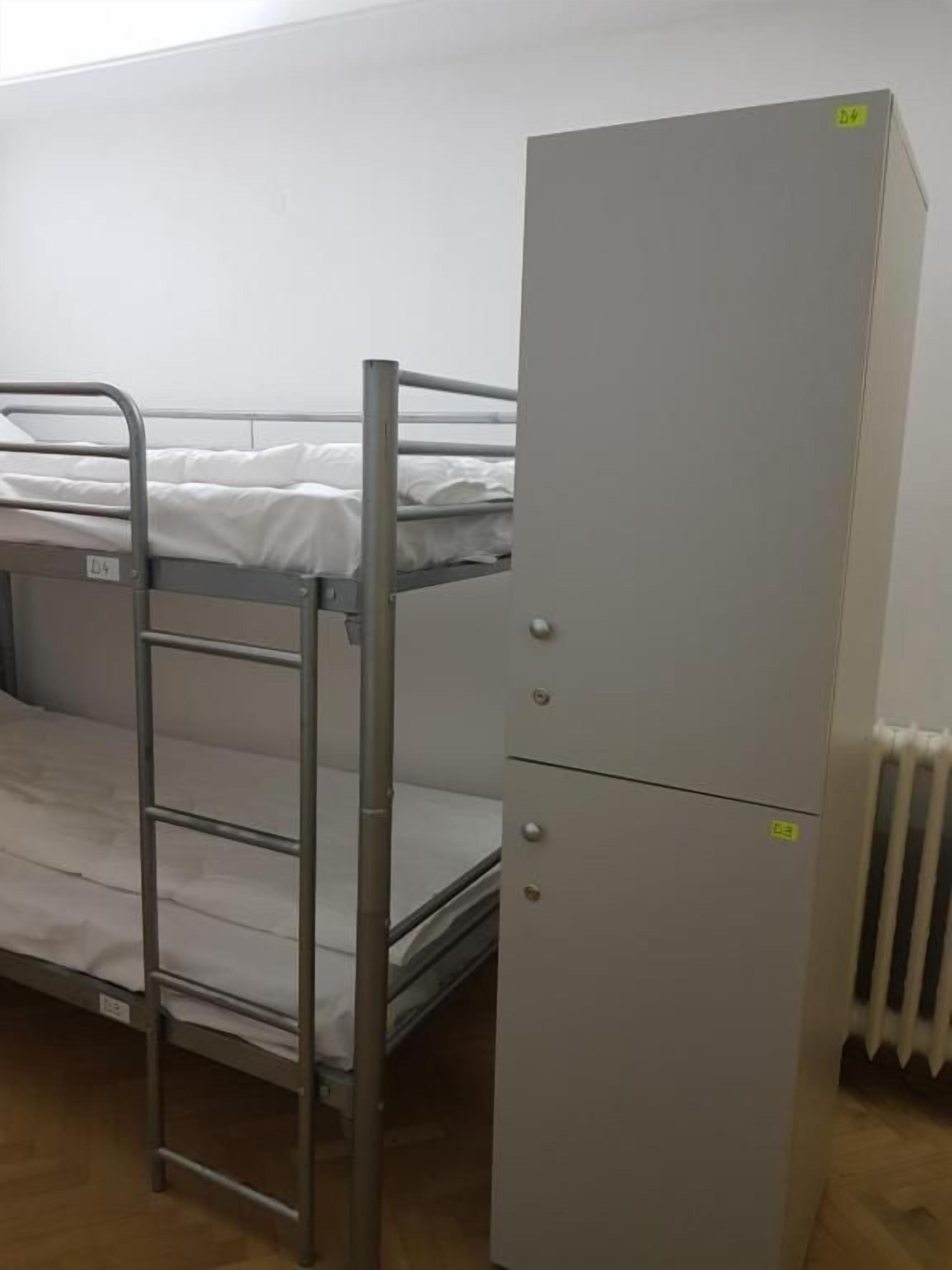 Una cama litera con escalera metálica, un armario pequeño y un radiador.