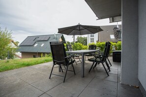 Eine Terrasse im Freien mit Tisch und Stühlen, einem großen Sonnenschirm und Blick auf ein Wohngebäude mit Satteldach.