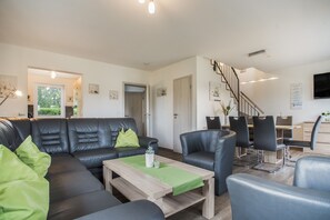 Una sala moderna con un sofá de cuero negro, cojines verdes, una mesa de centro de madera y un comedor con sillas negras y una mesa de madera.
