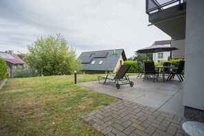 Eine Terrasse mit Rasenfläche, einem Liegestuhl, einem Tisch mit Stühlen und einem Gebäude mit geneigtem Dach.