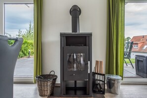 Una estufa de leña en una habitación con un ventanal grande, una cesta de mimbre y un cubo de metal.