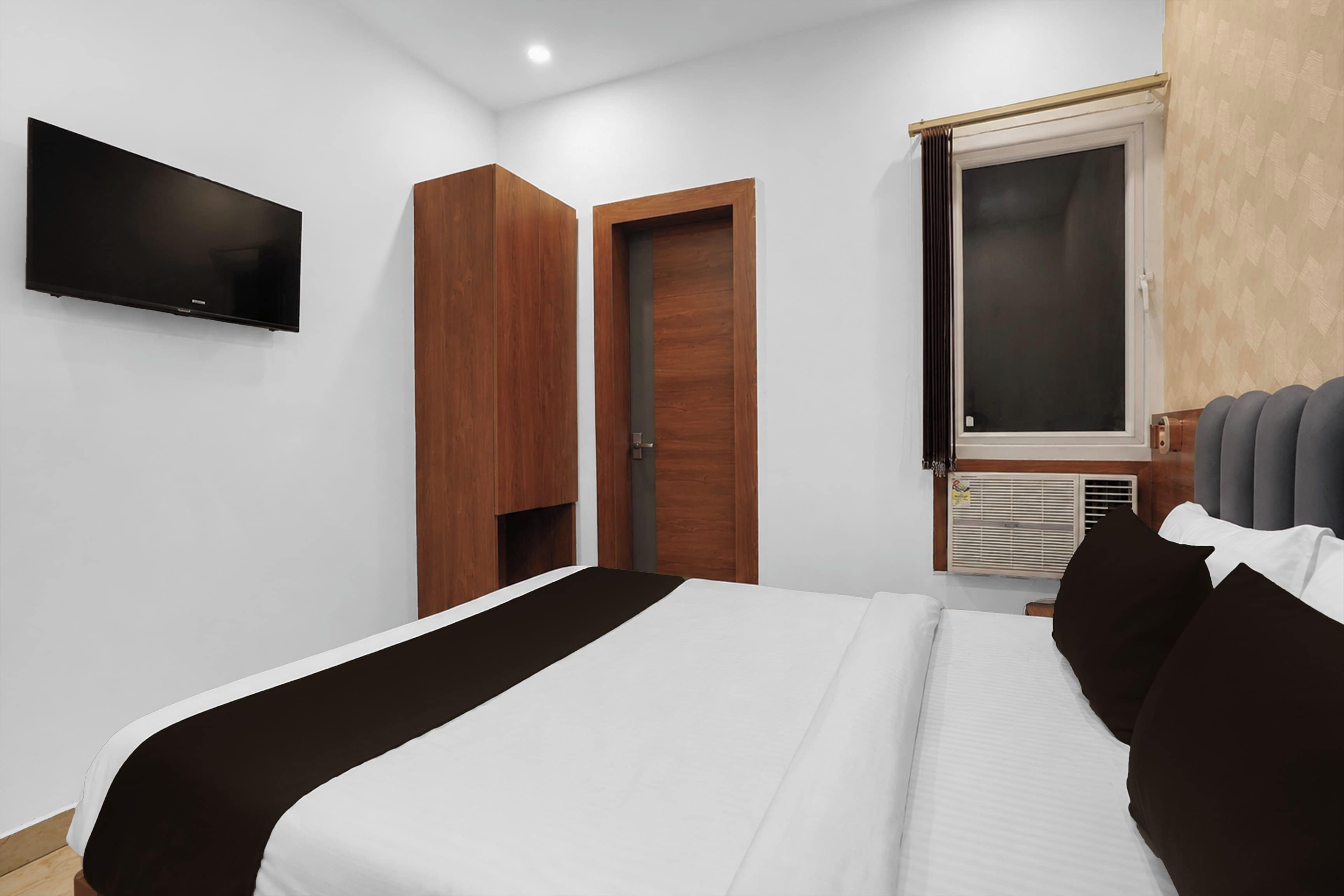 Una habitación de hotel con una cama, un televisor de pantalla plana montado en la pared, una ventana con persianas y un equipo de aire acondicionado incorporado.