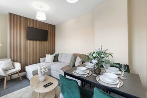 Een moderne woonkamer met een bank, een eettafel voor twee personen en een aan de muur bevestigde televisie.
