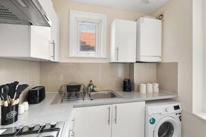 A modern kitchen with a built-in washing machine, a window with a view, and a sink with a dish rack. - Stylish Brick Lane Apartment Sleeps 9 Shoreditch (Greater London)